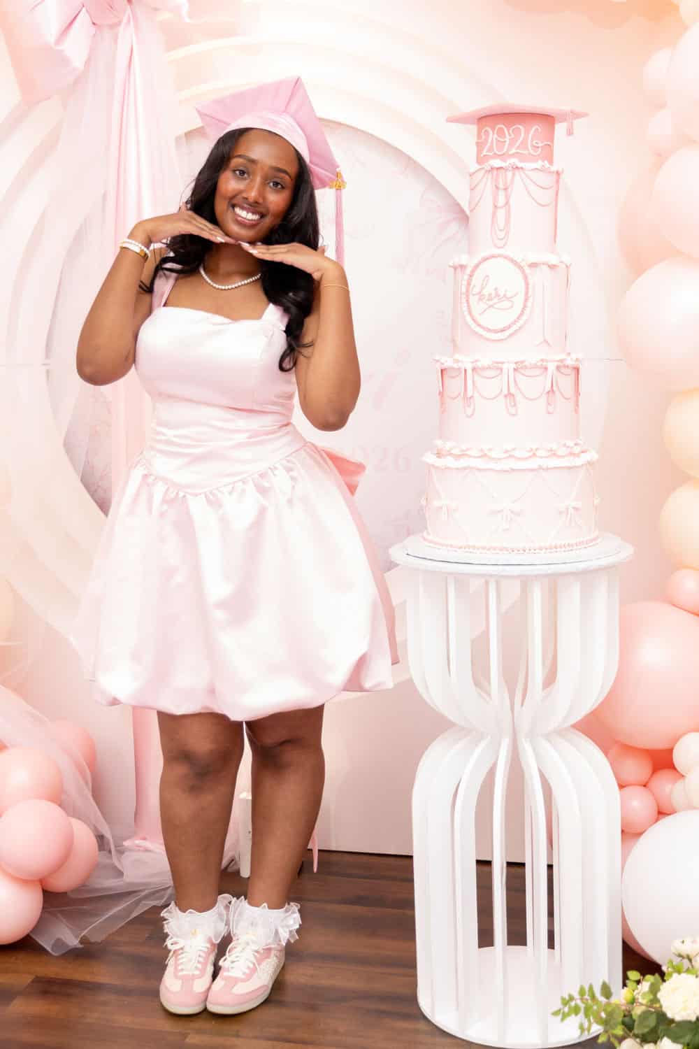 Young woman in pink dress and cap celebrating graduation with cake and balloons.