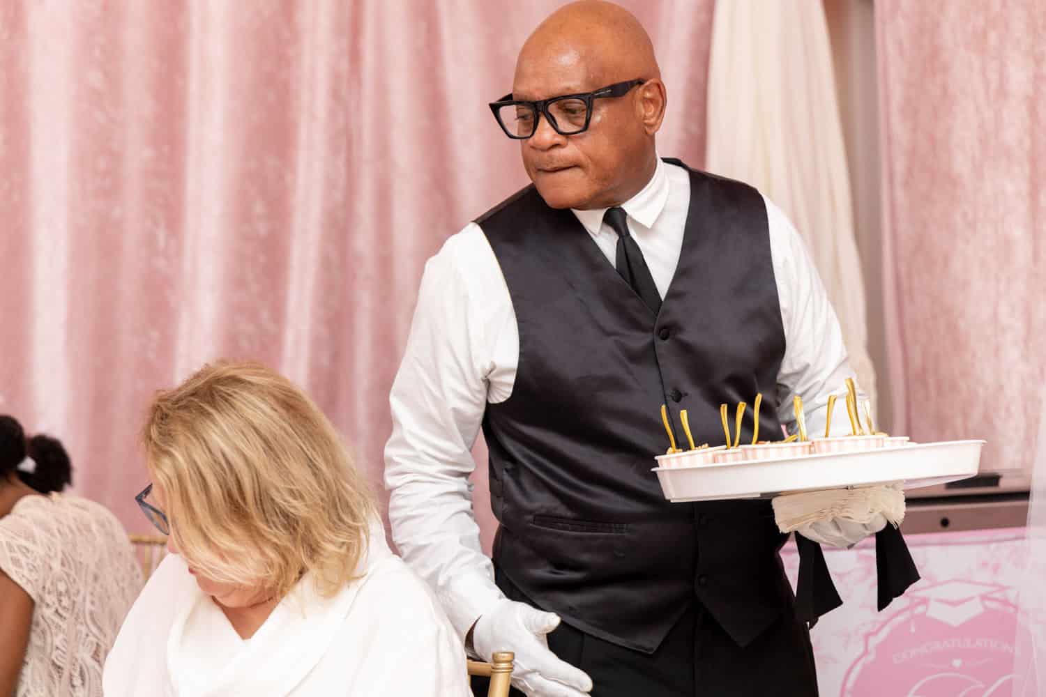 Professional man at event holding a cake with candles, wearing glasses and a vest.