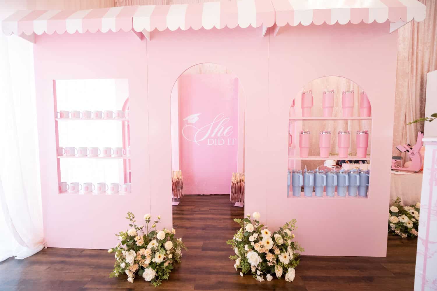 Decorative pink event display with floral arrangements and shelves of pink and blue bottles.