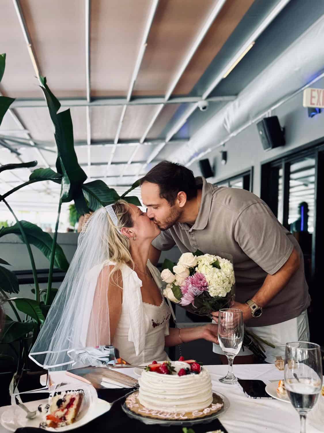 Bride and groom sharing a kiss at wedding reception with flowers and cake.