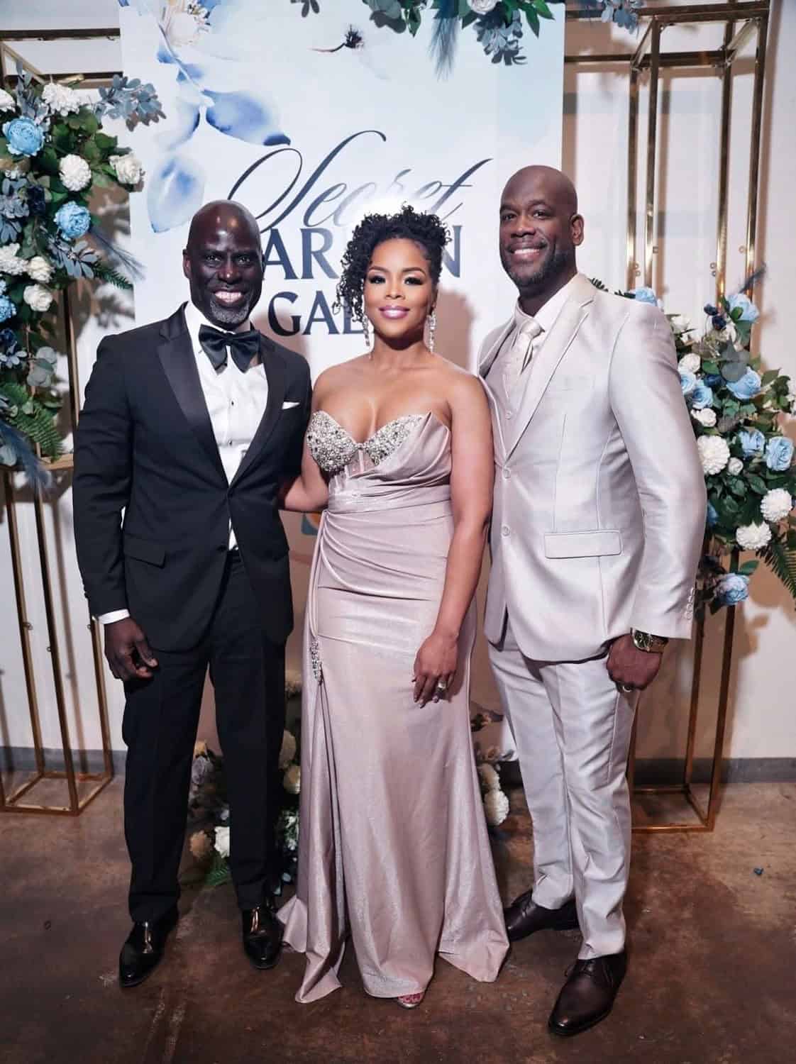 Three well-dressed individuals at a formal event with floral decor.