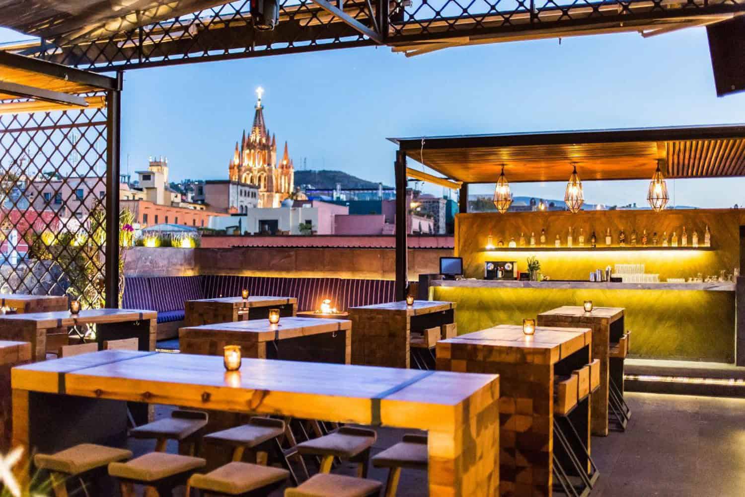 Rooftop bar overlooking city skyline with sunset sky and lit tables.