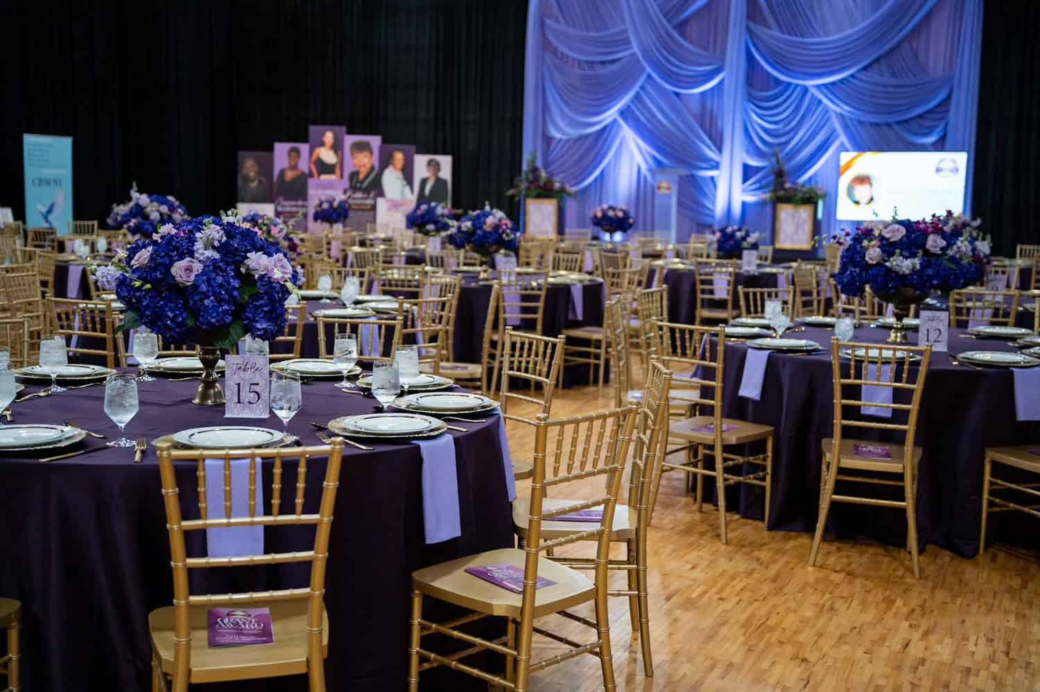 Event venue with round tables, purple tablecloths, and floral centerpieces.