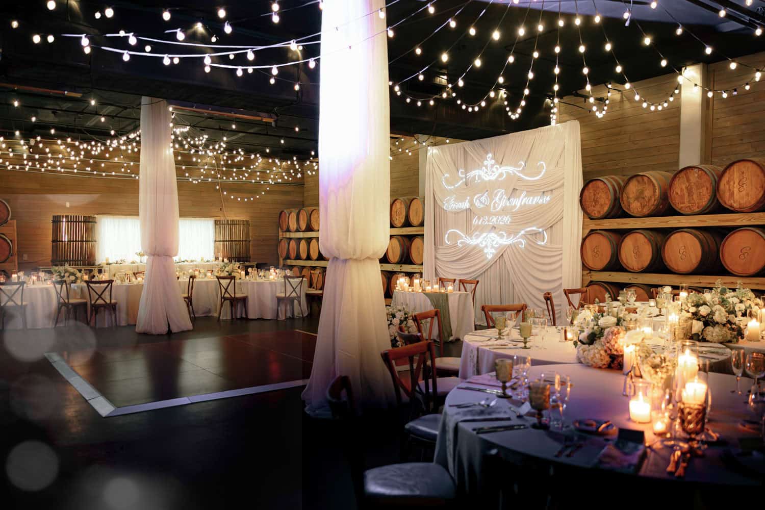 Indoor event space decorated with string lights, tables, and wine barrels for a celebration.