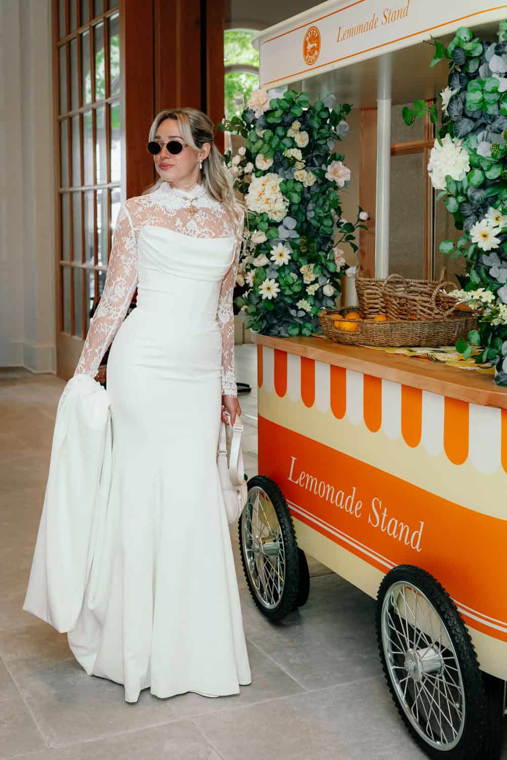 Stylish woman in white dress with lemonade stand at event.