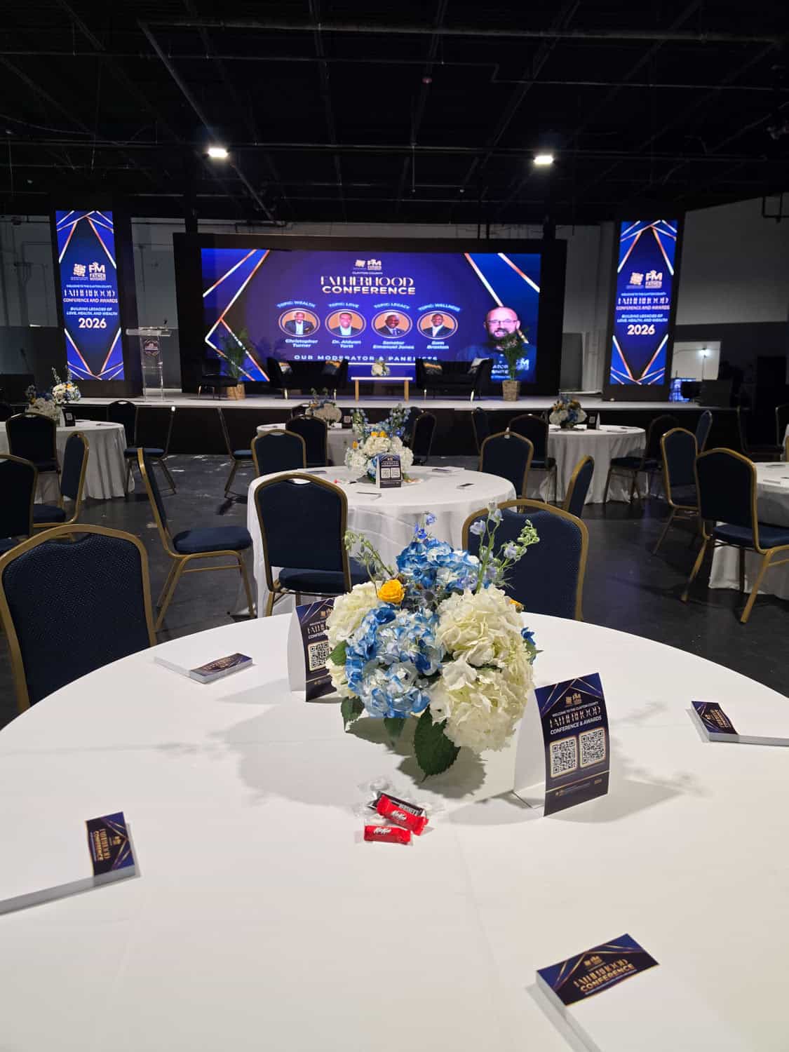 Conference room with round tables, floral centerpieces, and a large stage with screens.